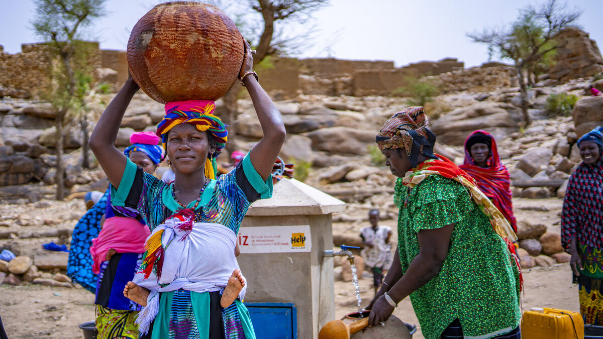 A Malian woman stands in front of a well and carries a container of water on her head
