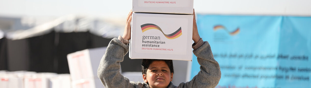 Ein syrischer Junge trägt zwei Hilfspakete mit der Aufschrift "German Humanitarian Assistance" auf seinem Kopf