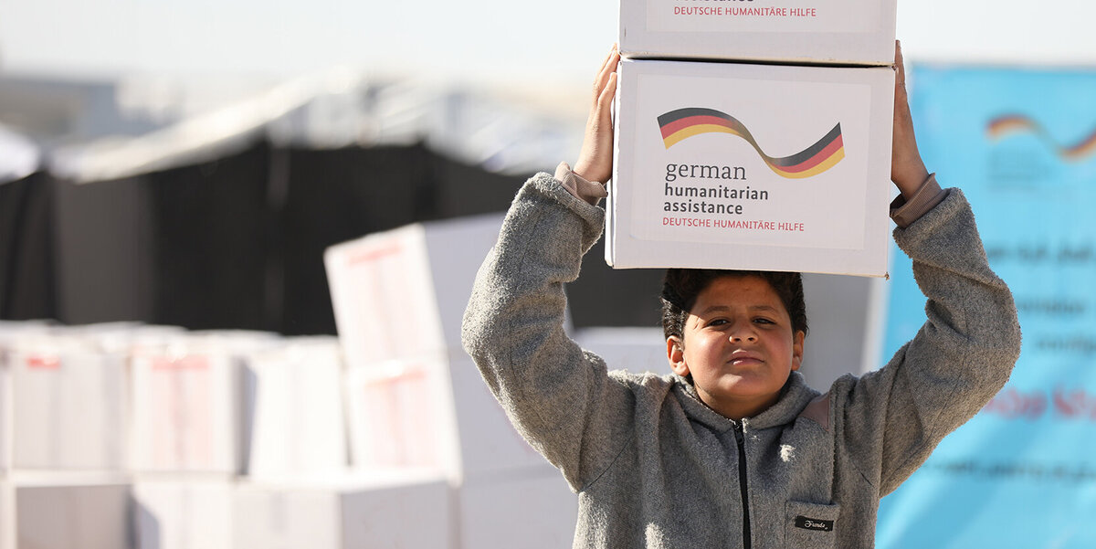 Ein syrischer Junge trägt zwei Hilfspakete mit der Aufschrift "German Humanitarian Assistance" auf seinem Kopf