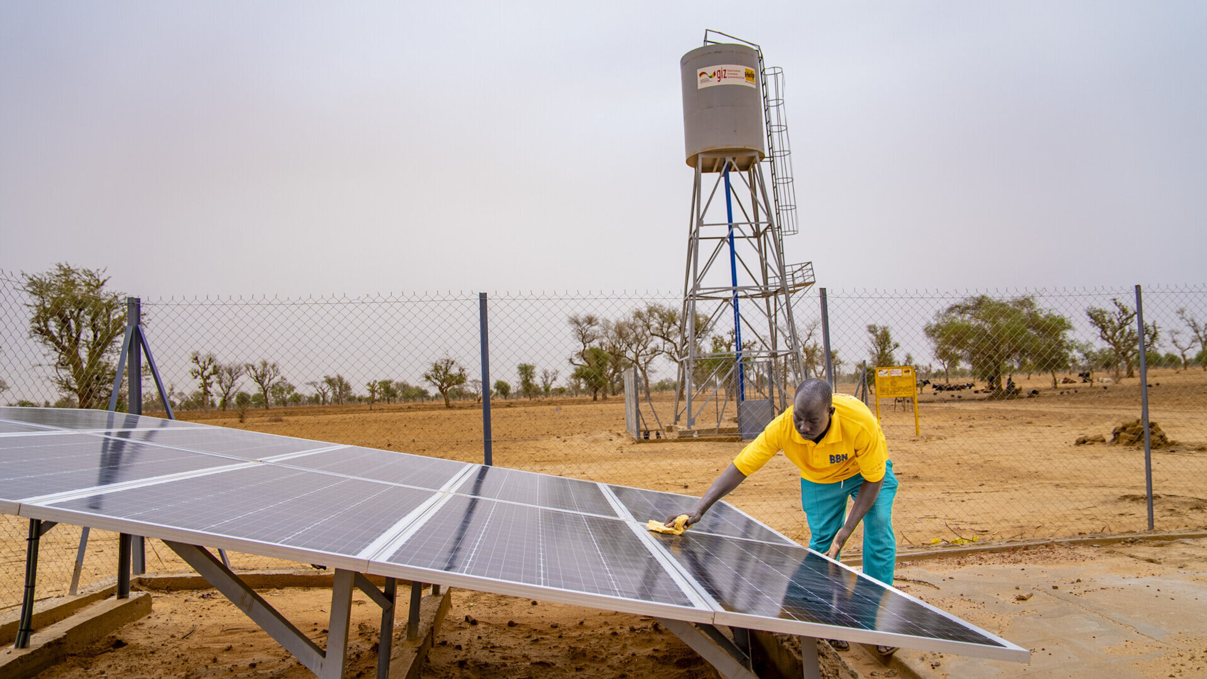A man cleans solar panels in a dry region, a water tower with the Help logo stands in the background