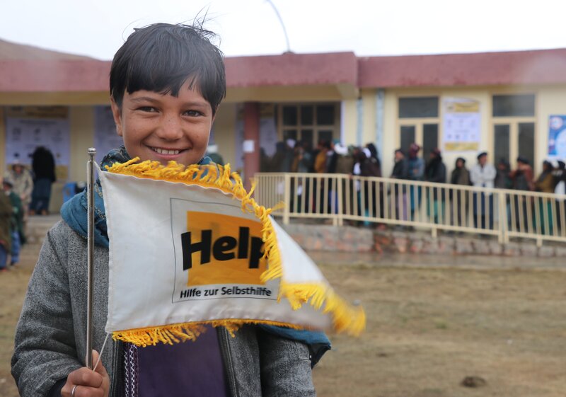 Ein afghanischer Junge hält eine Fahne mit dem Help-Logo in der Hand