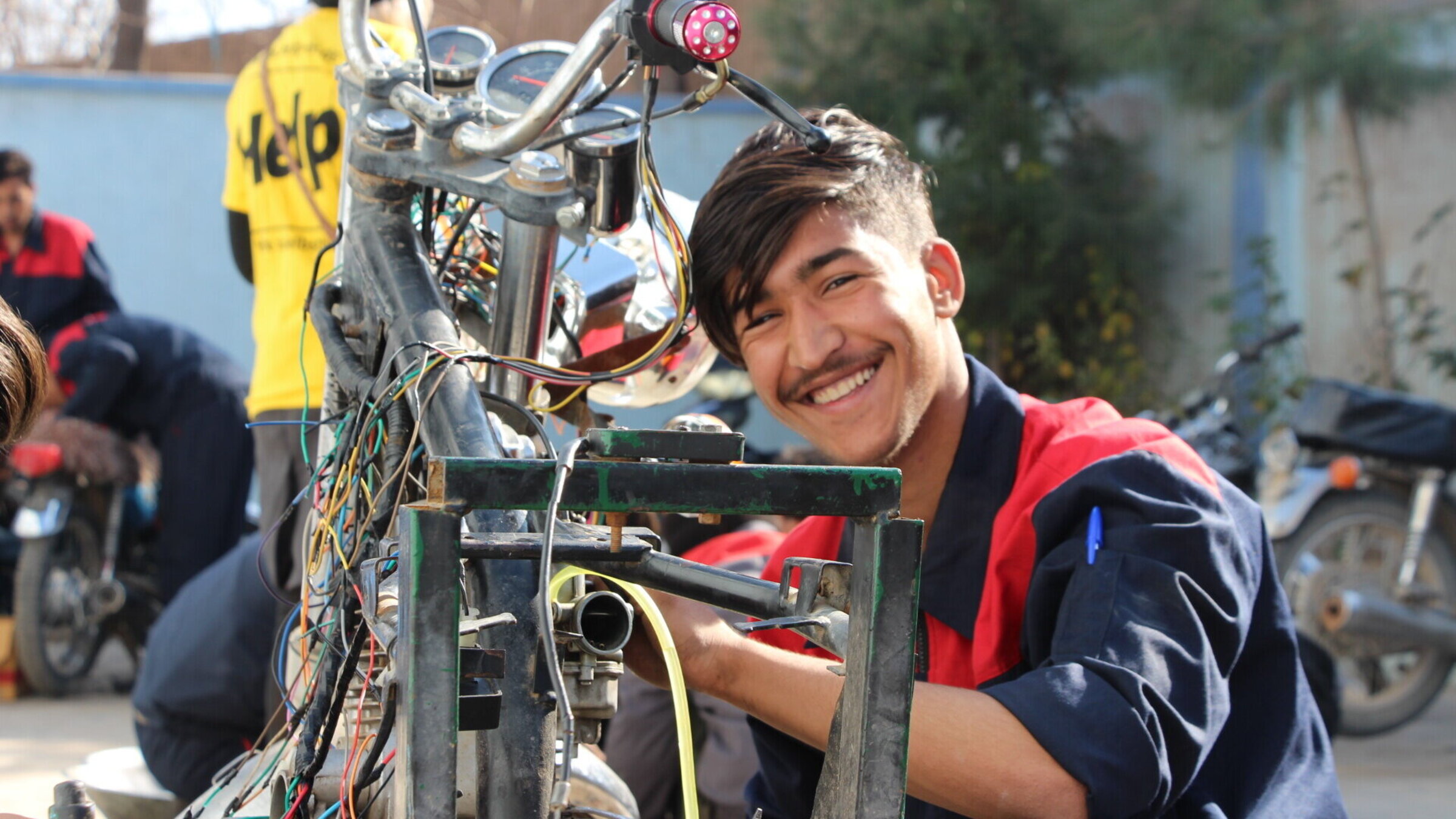 A young Afghan repairs a motorcycle and looks into the camera with a smile on his face