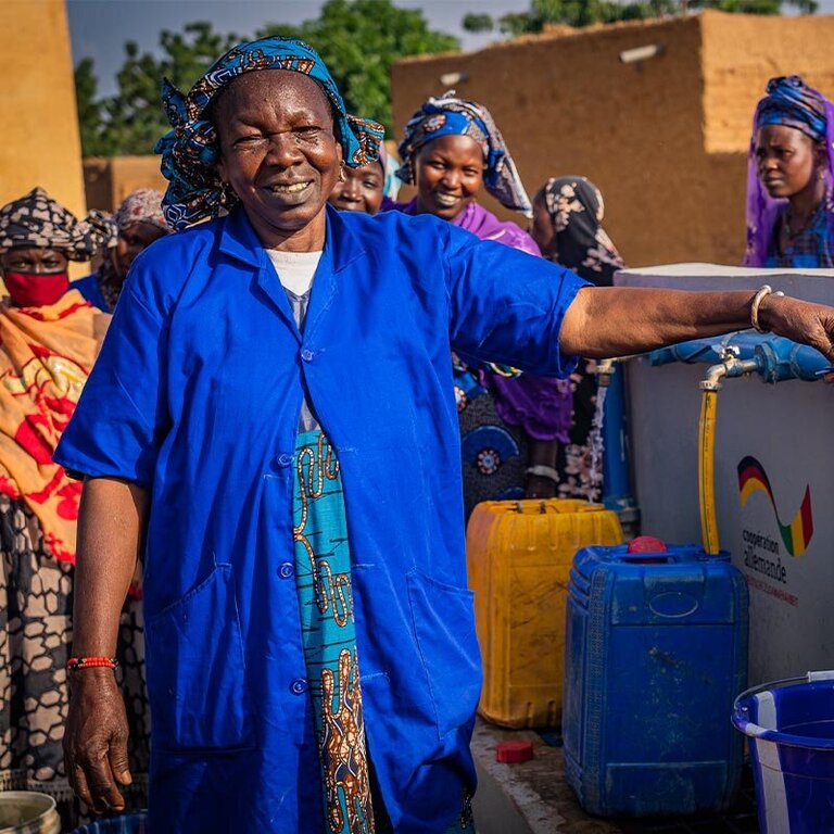 Spenden Mali: Help baut Trinkwasserbrunnen Eine Frau in einem blauen Kleid steht vor einem Help-Brunnen und zapft Wasser ab